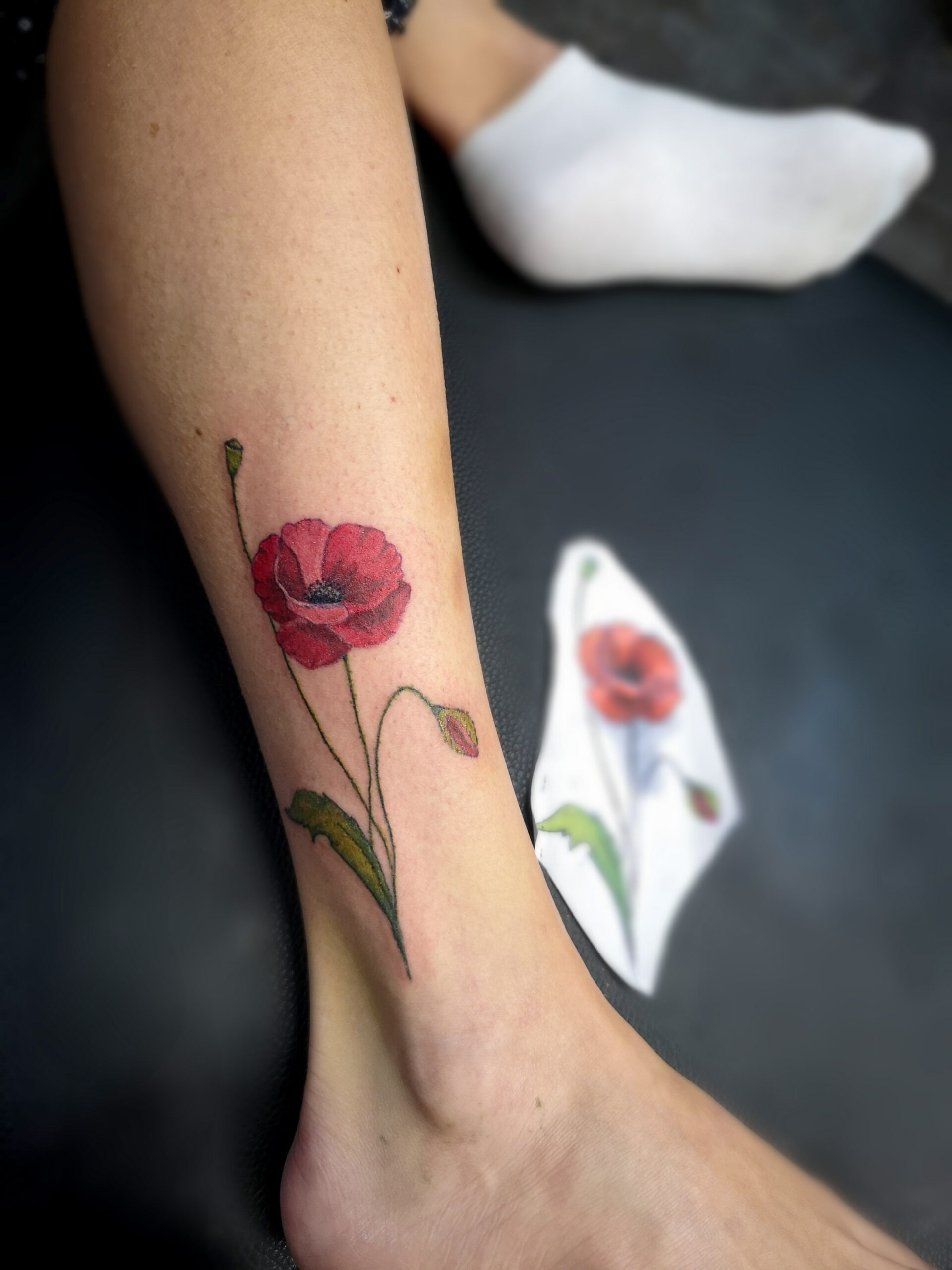 Photo d'un tatouage sur la cheville d'un coquelicot réaliste avec du rouge pour les pétales et du vert pour la tige