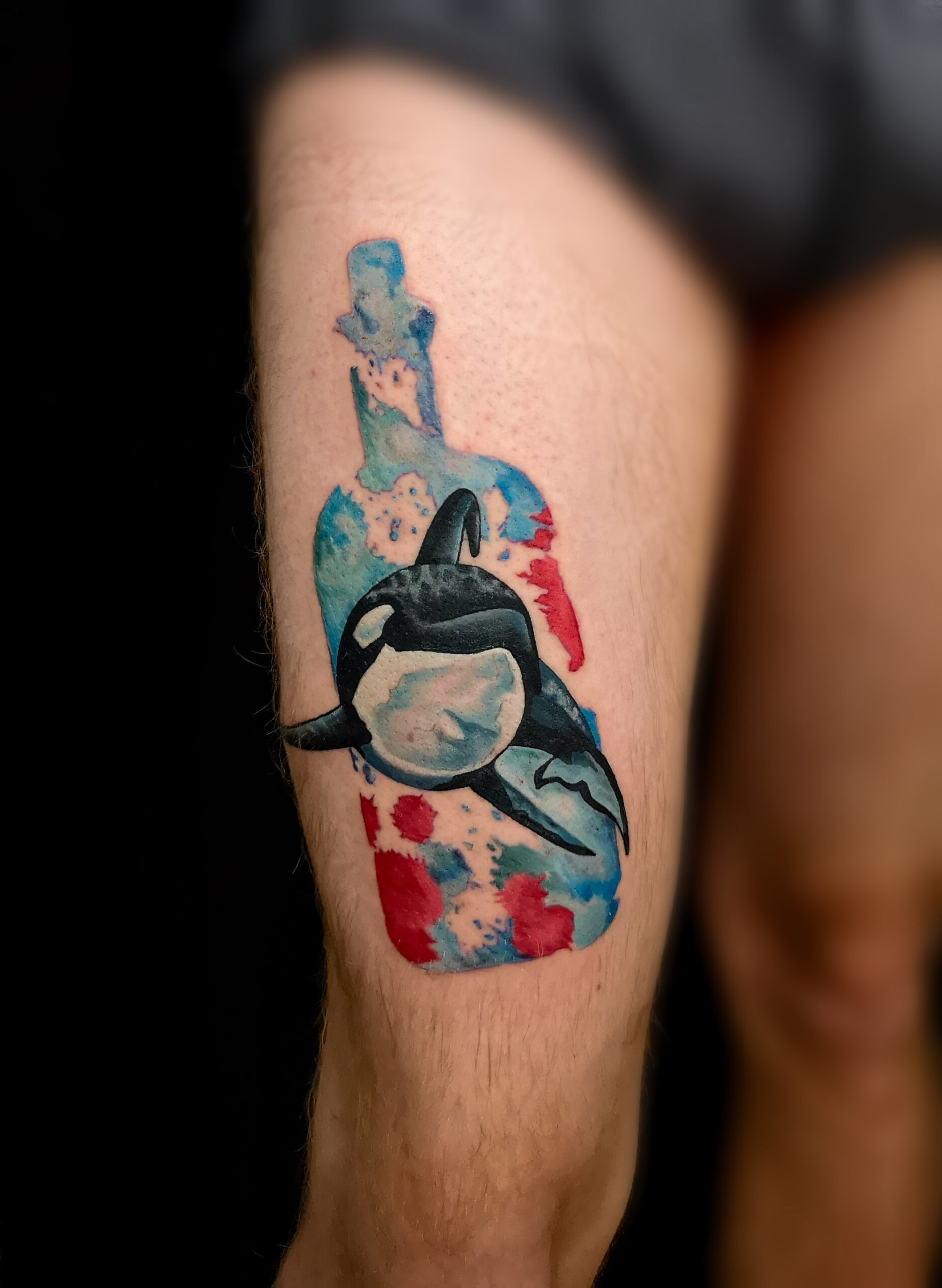 Photo d'un tatouage sur la cuisse d'une orque prisonnière dans une bouteille La bouteille est dans un style aquarelle en bleu et en rouge