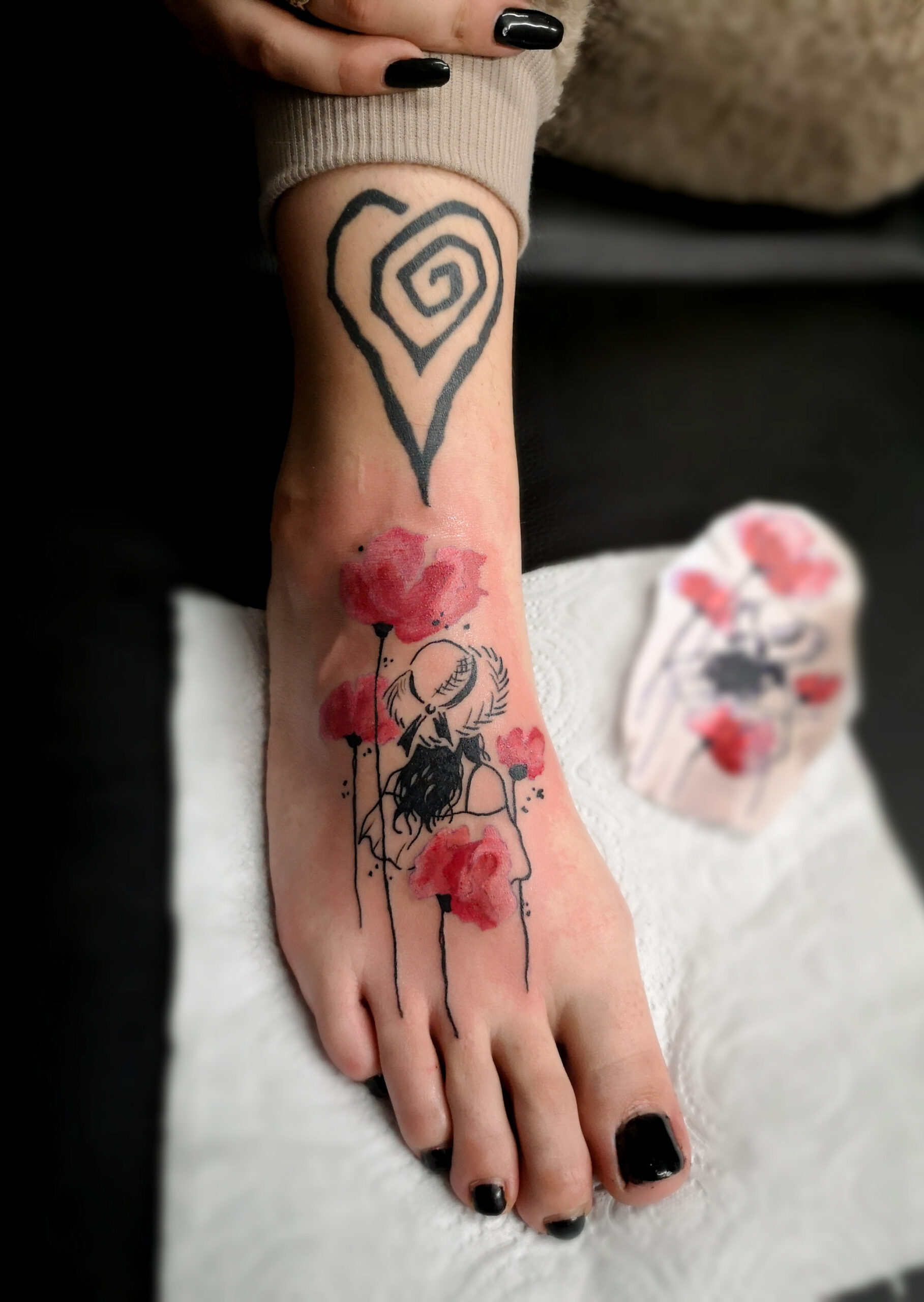Photo d'un tatouage sur le pied représentant une jeune fille de dos avec un chapeau de paille en style croquis et des coquelicots en aquarelle rouge et au dessus sur la cheville un cœur tourbillon