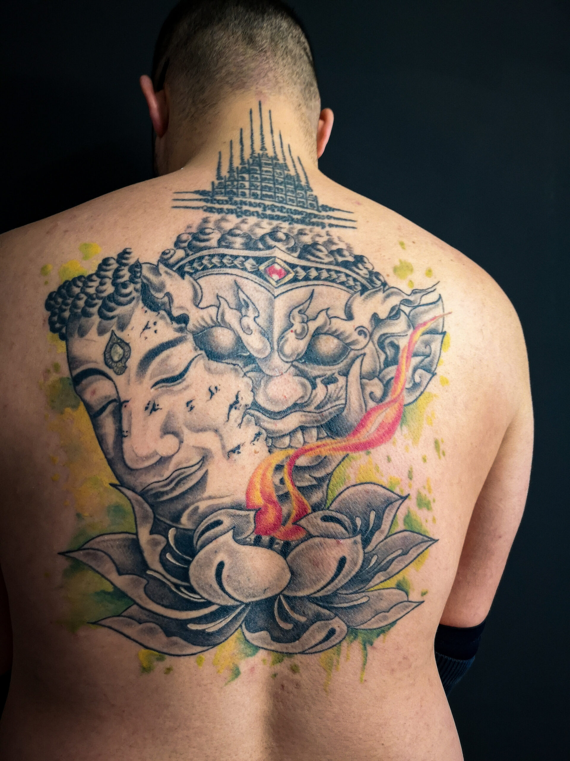 Photo d'un tatouage de pépère sur le dos représentant un masque de Bouddha sur un masque de démon au dessus d'un lotus en feu avec un fond aquarelle vert