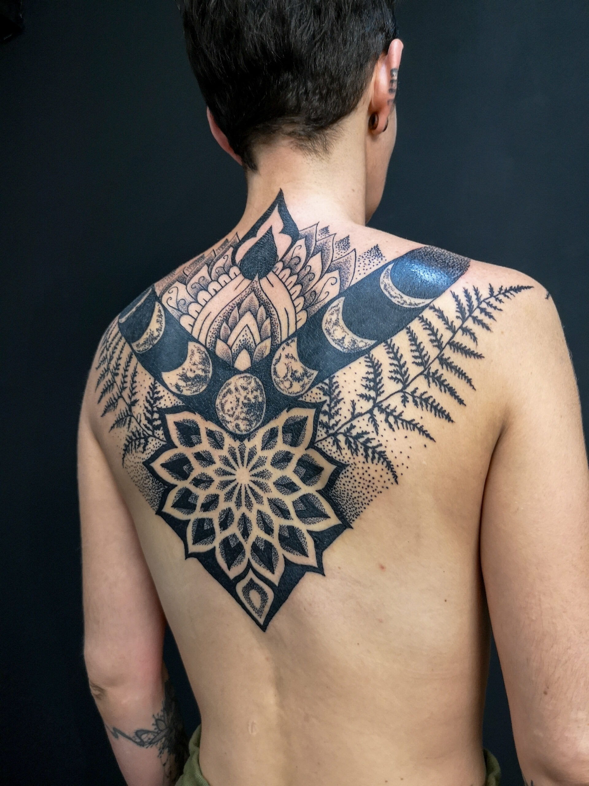 Photo d'un tatouage du dos dans un style graphique représentant les phases lunaires, un gros mandala et des feuilles de fougères