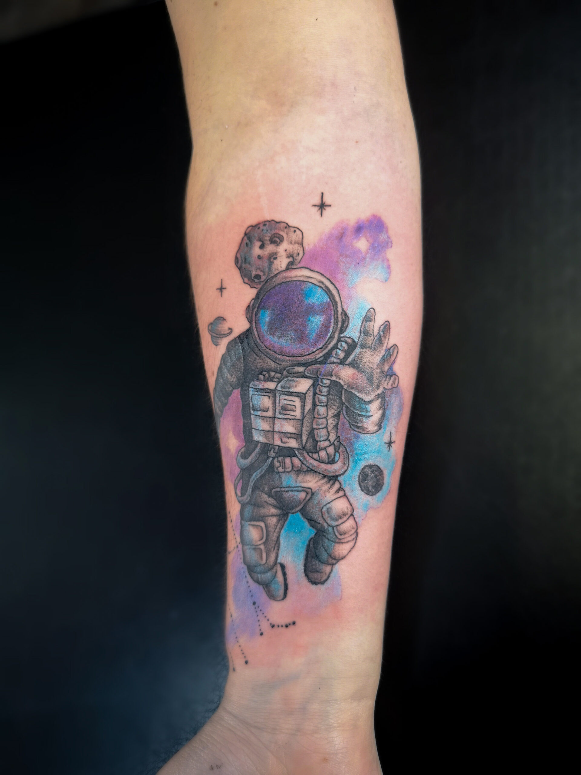 Photo d'un avant bras représentant un tatouage d'un cosmonaute dans l'espace avec en fond la galaxie en aquarelle bleu et violet