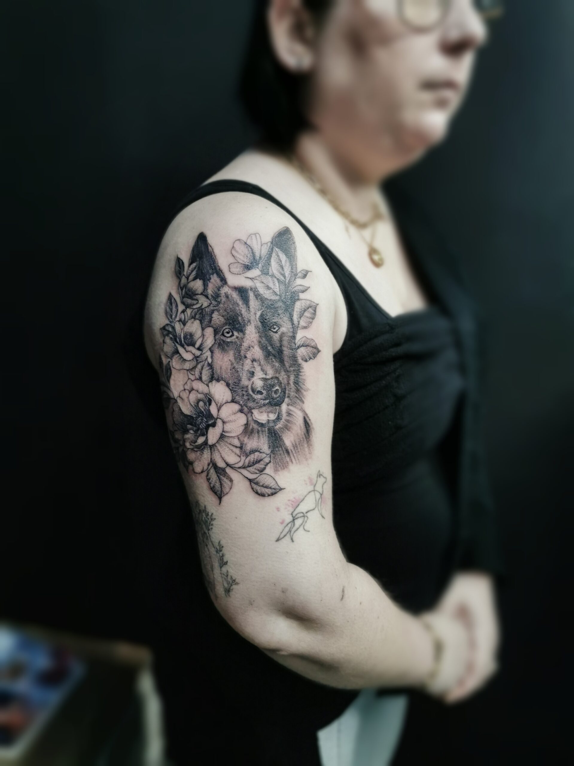 Photo d'un tatouage sur le haut du bras représentant le portrait d'un berger allemand entouré de fleur en style réaliste