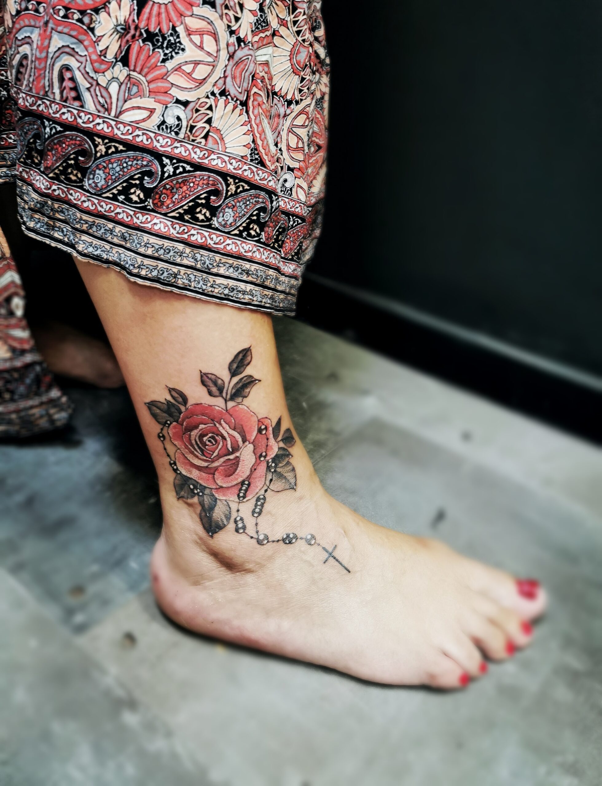 Photo d'un tatouage sur la cheville représentant une rose rouge et un chapelet dont la croix va sur le pied
