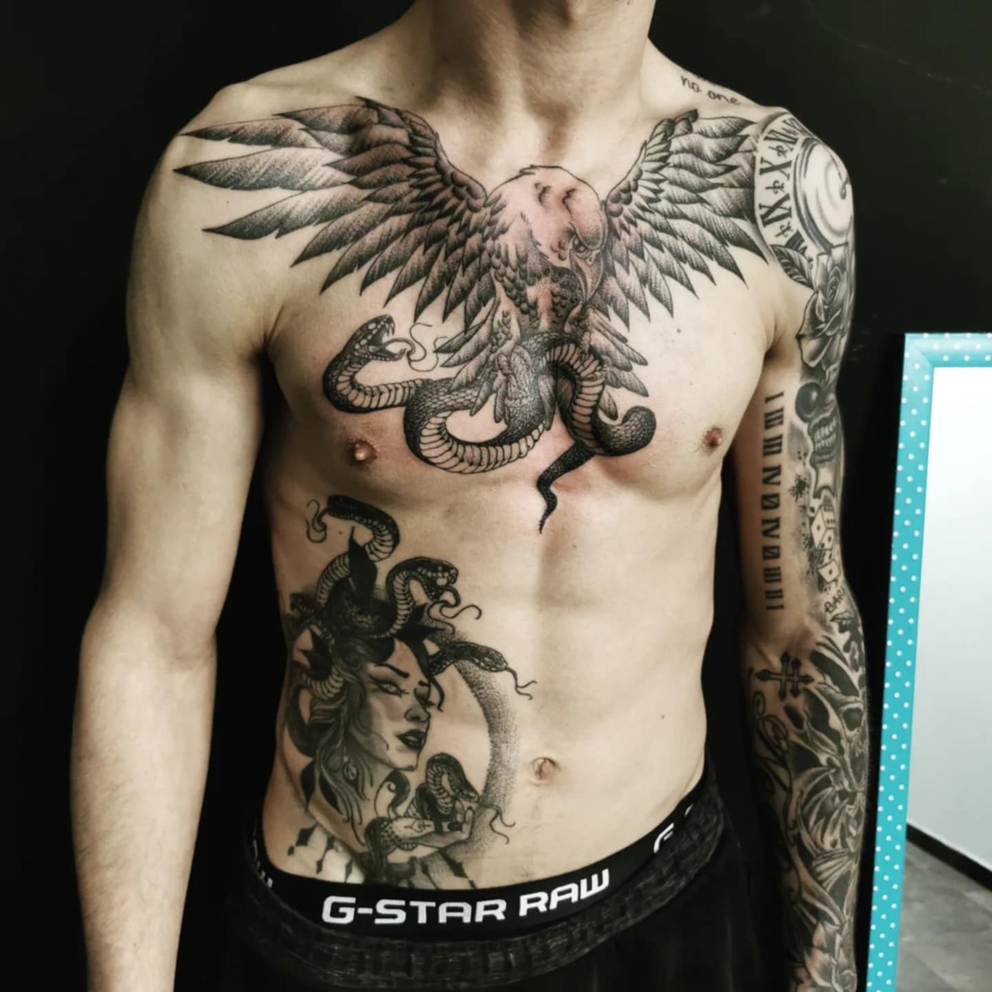 Photo d'un tatouage sur le torse , le ventre et le bras représentant respectivement un aigle tenant un serpent, un portrait de médusa et enfin du bio orga le tout en noir et gris